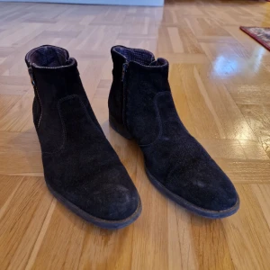 Svarta Playboy mockaboots med dragkedja - Snygga svarta Playboy boots i mocka med rund tå och dragkedja på sidan. Skorna har en låg klack och är fodrade med ett mönstrat innerfoder. Perfekta för höst och vinter när du vill ha en stilren look. Passar storlek 43/44