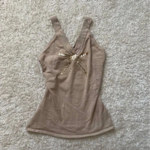 Beige linne med paljettrosett Saint Tropez - Supersött linne från Saint Tropez med breda axelband och v-ringning. Framtill sitter en stor rosett dekorerad med guldiga paljetter som ger extra bling. Perfekt för dig som vill sticka ut lite extra i sommarvärmen! Strl L men passar även S