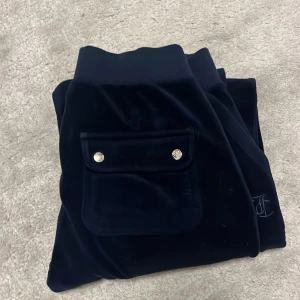 Mörkblåa mjukisbyxor i sammet Juicy Couture - Mörkablåa mjukisbyxor i sammet från Juicy Couture med bred resår och snörning i midjan. Byxorna har fickor fram och bak med lock och guldiga knappar samt broderad JC-logga framtill. Perfekta för en chill och snygg look.