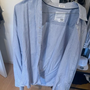 Blåvit randig skjorta från 157 - Klassisk skjorta med smala blå och vita ränder från 157. Skjortan har lång ärm, knappar framtill och en avslappnad passform. Perfekt för dig som gillar en stilren och enkel look. Tillverkad i mjuk bomull som känns skön mot huden.