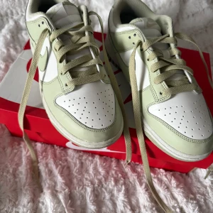 Nike Dunk Low i olive aura/sail-coconut milk - Nike Dunk Low sneakers i en fräsch ljusgrön och vit färgkombination. Skorna har klassisk snörning, rund tå och platt sula. Ovandelen är i slätt skinn med meshfoder för extra komfort. Perfekt för dig som gillar stilrena sneakers med en soft vibe.