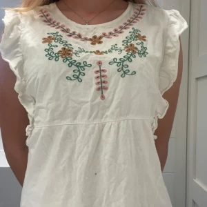 Vit broderad sommarblus med volang - Superfin vit blus med broderade blommor i grönt, rosa och gult vid halsen. Modellen är kort med volangdetaljer på axlarna och är ärmlös. Perfekt för varma dagar och har en avslappnad, bohemisk vibe. Materialet känns som bomull och blusen har en luftig passform.