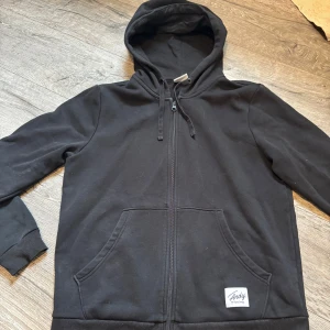 Svart hoodie från Andy by Frank Dandy - Svart hoodie med dragkedja och huva från Andy by Frank Dandy. Tröjan har snörning vid huvan, två stora fickor framtill och ribbade muddar vid ärmslut och nederkant. Perfekt för chill dagar och streetwear-stil.