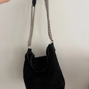 Svart axelväska med kedjerem - Säljer en svart axelväska i mocka med bred, mjuk form och coola silverfärgade kedjor som axelrem. Väskan är rymlig och har en stilren look som passar till många outfits. Perfekt för dig som gillar en edgy vibe och vill ha plats för allt du behöver. Passar perfekt nu till Skolstarten och hösten 🍁🍂