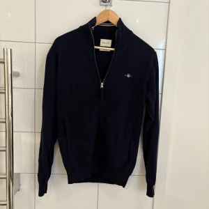 Mörkblå kofta från GANT i lammull - Snygg mörkblå kofta från GANT i superfin lammull. Tröjan har hel dragkedja framtill, ribbade muddar och diskret GANT-logo på bröstet. Perfekt för dig som gillar klassisk stil med en modern touch. Passar dig som vill ha en varm och mjuk tröja med lång ärm.