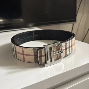 Burberry bälte  - Säljer ett Burberry bälte med ikoniskt TB-spänne i silver. Ena sidan har klassiskt beige-rutigt Burberry-mönster, andra sidan är svart med diskret rutmönster och ingraverad ryttare. Bältet är i skinn och kan vändas för olika looks. Perfekt accessoar för att lyfta din outfit.