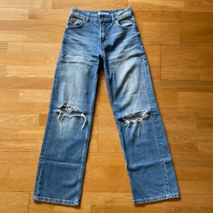 Bershka 90s wide jeans med slitningar - Blåa 90-talsinspirerade wide jeans från Bershka med hög midja och raka ben. Snygga slitningar över knäna och klassisk femficksmodell. Jeansen är i denim och har en cool, avslappnad vibe.