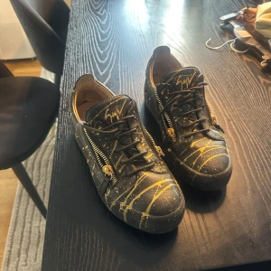 Svarta sneakers från Giuseppe Zanotti - Unika äkta svarta sneakers från Giuseppe Zanotti med guldiga stänk och detaljer. Skorna har snörning, dragkedjor på sidorna och signaturlogga på plösen. Tillverkade i skinn med cool konstnärlig vibe och platt sula. Perfekta för dig som vill sticka ut. 