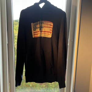 Mörkblå hoodie Ralph Lauren flag patch - Snygg mörkblå hoodie från Ralph Lauren Denim & Supply med stor amerikansk flag patch på bröstet. Tröjan har huva med snörning och känguruficka framtill. Tillverkad i 100% bomull för skön känsla och passform.