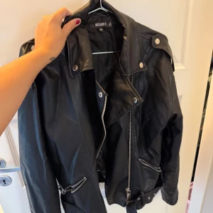 Svart skinnjacka från Missguided - Säljer en svart skinnjacka från Missguided med coola silverfärgade dragkedjor, knappar och bälte i midjan. Storlek 34 men är lite oversize.