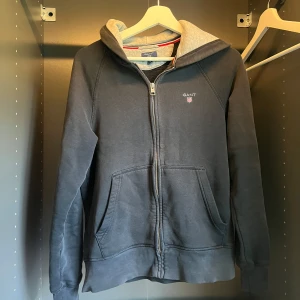 Svart hoodie från GANT med dragkedja - Snygg svart hoodie från GANT i bomull med dragkedja framtill och broderad logga på bröstet. Tröjan har huva med ljusgrå insida och två stora fickor framtill. Perfekt för en chill och avslappnad stil. Passar stolek S. lite solblekt