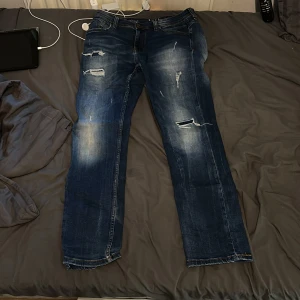 Mörkblå slitna jeans från Smog - Mörkblå jeans från Smog med coola slitningar och distressed detaljer på benen. Klassisk femficksmodell med raka ben och kontrastsömmar. Jeansen har en snygg tvätt med ljusare partier och är tillverkade i bomull för en skön känsla. Storlek 30/30/M sparsamt använd. 