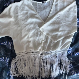 Vit blus med fransar från H&M - Superfin vit blus från H&M med v-ringning, knappar framtill och långa fransar längs nederkanten. Blusen har trekvartsärm och är gjord i ett lätt, flowy material som känns riktigt skönt. Perfekt för dig som vill sticka ut med coola detaljer!
