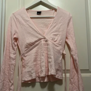 Ljusrosa omlottopp från Gina Tricot - Supersöt ljusrosa omlottopp från Gina Tricot i storlek XS. Toppen har långa ärmar, v-ringning och knytband i midjan. Den har aldrig används och bara legat i min garderob så därför är den lite skrynklig!