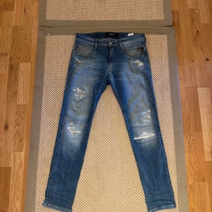 Blå Replay jeans med slitningar - Säljer ett par blå Replay jeans med snygga slitningar och distressed detaljer på benen. Jeansen har klassisk femficksdesign, normal passform och är tillverkade i mjukt denimtyg. Perfekta för en avslappnad och trendig look.