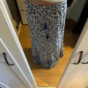 Mönstrad långkjol från Vero Moda - Supersöt långkjol från Vero Moda i vitt och blått med blommigt mönster. Kjolen har resår i midjan och snörning med tofsar framtill. Bara andvänd en gång så i väldigt bra skick. Går hela vägen ner till marken på mig som är 178cm🌸🌸🌸🌸🌸
