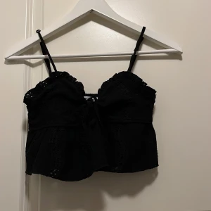 Svart spetsig linnetopp FB Sister - Supersöt svart linnetopp från FB Sister med smala axelband, volangkant och dekorativ snörning framtill. Toppen har fina spetsdetaljer och broderade partier som ger en romantisk vibe. Perfekt att styla med jeans eller kjol för en trendig look.