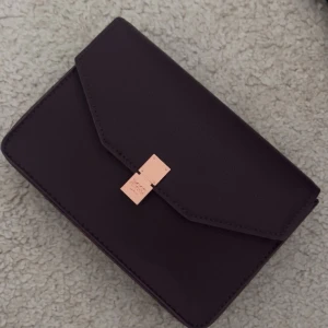 Lila handväska från hugo boss - Säljer en stilren lila handväska från hugo boss med guldfärgat spänne framtill. Väskan är i slätt skinn och har en enkel, boxig form med lock. Perfekt storlek för det viktigaste och en snygg detalj till din outfit.