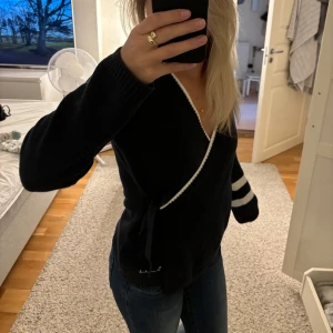 Svart omlott kofta med vita detaljer - Snygg svart omlott kofta med vita ränder på ena ärmen och vit kant längs den v-ringade halsen. Koftan har knyt i sidan och är långärmad.