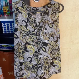 Mönstrad ärmlös blus från H&M - Säljer en ärmlös blus från H&M med paisleyinspirerat mönster i svart, vitt, gult och brunt. Blusen har en lös passform och liten v-ringning framtill. Perfekt för dig som gillar färgstarka och detaljerade prints.