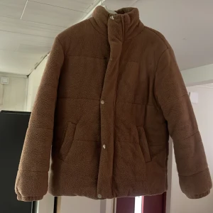 Brun teddyjacka med dragkedja - Mysig brun teddyjacka med hög krage och dragkedja framtill. Jackan har två snedställda fickor och stängs med både dragkedja och tryckknappar. Perfekt för kalla dagar när du vill vara både varm och snygg.