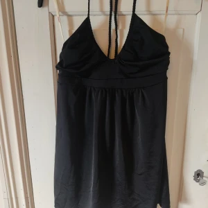 Svart topp med flätade band - En svart topp/tunika med flätade band som går att knyta som man vill runt nacken så den sitter som en halter. Tröjan är av ett lätt glansigt tyg med mycket stretch. 