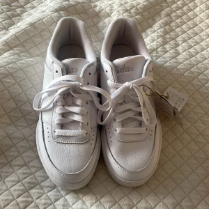 Vita Adidas sneakers i skinn - Fräscha helvita Adidas sneakers i skinn med klassisk låg siluett och rund tå. Skorna har snörning och diskret Adidas-logga på plösen. Snygg och stilren modell som passar till allt, levereras i originalkartong. Aldrig använda. 