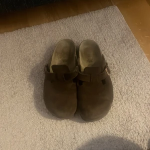 Birkenstock Boston mocka brun - Säljer ett par Birkenstock Boston i brun mocka med justerbart spänne i metall. Skorna har en klassisk rund tå och platt sula, perfekt för chill dagar. Materialet är mjukt och ger en skön känsla. Ikonisk slip-on modell som är enkel att ta på och av.