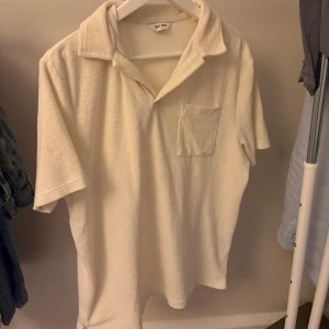 Vit piké tröja - Vit t-shirt från East West i frottématerial med krage och bröstficka. Modellen är kortärmad och har en loose passform som ger chill vibe. Perfekt för dig som gillar mjuka och sköna plagg med lite retro känsla.