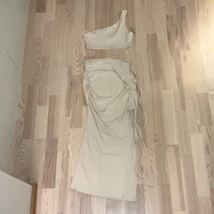 Beige set med kjol och one shoulder topp  - Säljer en beige ribbad topp med one shoulder-design och tillhörande kjol med en slits. Toppen är croppad och har en stilren look. Materialet är mjukt och stretchigt, vilket gör den bekväm att bära. Enkel men trendig modell för dig som gillar snygg stil.