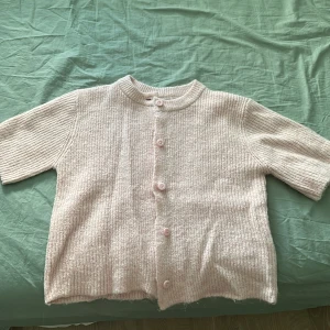 Ljusrosa stickad kofta från Gina Tricot - Säljer en söt ljusrosa stickad kofta från Gina Tricot med korta ärmar och rund hals. Koftan har ribbad struktur och knäpps med fem matchande knappar framtill. Perfekt att slänga över en topp för en chill look.