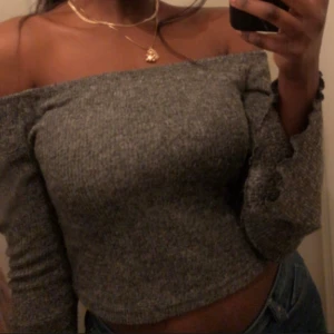 Grå offshoulder croppad stickad topp - Säljer en grå stickad offshoulder topp med lång ärm och croppad passform. Toppen har en ribbad struktur och sitter snyggt över axlarna. Perfekt att styla med jeans för en chill men trendig look.