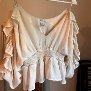 Beige/babyrosa volangblus från NLY Design - Supersöt beige/babyrosa blus från NLY Design med v-ringning, volangdetaljer längs ärmarna och smockad midja. Luftigt material och kortare modell som ger en romantisk vibe. Tröjan har även öppna ärmar! Perfekt att styla med jeans eller kjol för en trendig look. Den är ganska kort men så otroligt söt!💕