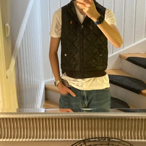 Svart väst från Ralph Lauren - Säljer en svart quiltad väst från Polo Ralph Lauren med orange logga på bröstet. Västen har dragkedja framtill, två fickor med tryckknappar och ribbade muddar nedtill. Perfekt att slänga över en hoodie eller t-shirt för en chill look.
