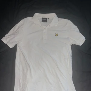 Vit pikétröja från Lyle & Scott - Säljer en klassisk vit pikétröja från Lyle & Scott med korta ärmar och krage. Tröjan har knäppning framtill och den ikoniska gula loggan broderad på bröstet. Perfekt till jeans eller shorts för en clean och stilren look.