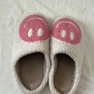 Vita tofflor med rosa smiley - Mysiga vita tofflor med fluffigt tyg och stora rosa smiley-motiv framtill. Perfekta att glida runt i hemma, med mjuk insida och vit sula. Tofflorna har en enkel slip-on design och är riktigt sköna för chill dagar. Storlek 38-39, osäker vart den är köpt ifrån men kommer inte mer till användning. Kontakta gärna innan köp!