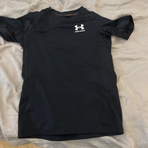 Svart Under Armour drift t shirt - Svart t-shirt från Under Armour med vit logga på bröstet. Modellen är kortärmad och har en tight passform, perfekt för träning. Tillverkad i syntetmaterial med HeatGear-teknologi som håller dig sval och bekväm under intensiva pass. Storleken är lite oklar men passar någon på 140-150cm lång skulle jag säga. FASTPRIS 89kr.