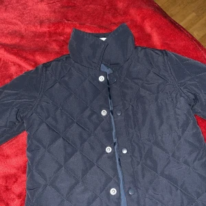 Quiltad mörkblå jacka från H&M - Säljer en quiltad mörkblå jacka från H&M med hög krage och tryckknappar framtill. Jackan har ett rutigt mönster i sömmarna och en bröstficka. Perfekt för kyliga dagar och enkel att styla till olika outfits.