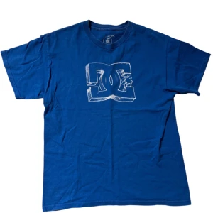 Blå t-shirt från DC - Säljer en blå t-shirt från DC med ett stort vitt logotryck på framsidan. T-shirten är tillverkad i 100% bomull och har en klassisk passform. Perfekt för en avslappnad stil.