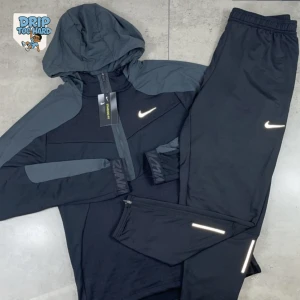 Svart Nike hoodie med huva - Svart hoodie från Nike med grå detaljer på axlar och ärmar. Tröjan har huva, dragkedja framtill och Nike-logga på bröstet. Perfekt för träning eller chill, med sportig look och bekväm passform.
