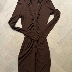 Brun skjortklänning med volangdetalj - Säljer en brun skjortklänning med långa ärmar och knappar hela vägen framtill. Klänningen har en snygg krage och volangdetaljer längs knappraden. Materialet känns mjukt och stretchigt, perfekt för dig som gillar en figurnära passform. I nytt skick