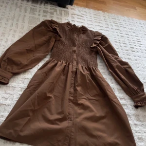 Brun klänning med volang och knappar - Säljer en brun klänning med lång ärm, volangdetaljer vid axlarna och smockad överdel. Klänningen har knappar hela vägen framtill och är gjord i ett mjukt bomullsmaterial. Perfekt för dig som gillar romantiska detaljer och en avslappnad passform.
