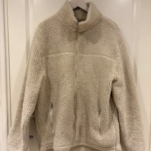 Beige teddyjacka från 157, storlek M - Mysig beige teddyjacka från 157 i storlek M. Jackan har hög krage, dragkedja framtill och två fickor med dragkedja. Perfekt för kyliga dagar när du vill vara både varm och stilren. Fleece-liknande material som känns supermjukt.