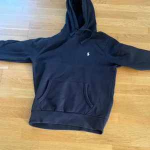 Mörkblå hoodie från Polo Ralph Lauren strl m - Snygg blå (färgen står som FADED BLACK) hoodie från Polo Ralph Lauren med klassisk broderad logga på bröstet. Tröjan har huva med snörning, stor magficka och långa ärmar. Ett litet hål på armbågen som är lagat. Väldigt sparsamt använd. 