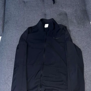 Svart vindjacka från Nike - Svart vindjacka från Nike med diskret logga på bröstet. Jackan har dragkedja framtill, ståkrage och är tillverkad i lätt polyester. Perfekt för dig som gillar sportig och clean stil. Långärmad modell med normal passform.