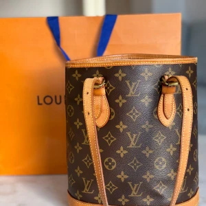 Louis Vuitton Monogram handväska - Säljer en klassisk Louis Vuitton handväska i brunt monogrammönster med LV-logga och blommor. Väskan har rund form, två smala handtag och detaljer i ljust skinn. Insidan har dragkedjefack och präglad Louis Vuitton-text. Perfekt för dig som gillar lyxiga accessoarer.
