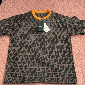 Fendi brun t-shirt med FF-mönster - Helt ny oanvändt snygg brun t-shirt från Fendi med det klassiska FF-logomönstret över hela plagget. Rund hals med kontrasterande gul kant ger en cool touch. Tillverkad i 100% bomull för skön känsla. Perfekt för dig som gillar lyxiga streetwear-vibbar.