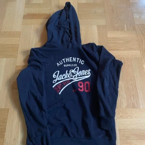 Mörkblå hoodie Jack & Jones - Säljer en mörkblå hoodie från Jack & Jones med tryck i vitt och rött på bröstet. Tröjan har huva med snörning och långa ärmar. Perfekt för chill dagar eller när du vill ha en avslappnad stil.