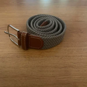 Grått flätat bälte med bruna detaljer - Snyggt grått flätat bälte med bruna skinn-detaljer och silverspänne. Perfekt accessoar för att lyfta din outfit med en stilren och modern touch. Bältet har en bred, vävd design som ger en cool struktur.