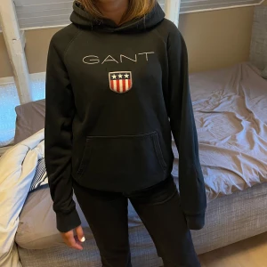 Svart hoodie från GANT med logga - Svart hoodie från GANT med broderad logga och sköld på bröstet. Använts några gånger. Klassisk känguruficka framtill och huva. Perfekt för dig som gillar stilren och sportig look. Mjuk och skön bomullsmix som passar till många outfits.